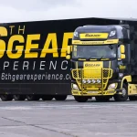 A black and yellow truck with the branding "6th Gear Experience" is parked on a paved surface. The truck has prominent yellow detailing on the front grille and bumper, with additional lights mounted on the front. The side of the truck features large yellow text reading "6th GEAR EXPERIENCE" and the website "www.6thgearexperience.com". The background shows a grassy field and some trees under an overcast sky.