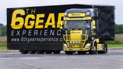 A black and yellow truck with the branding "6th Gear Experience" is parked on a paved surface. The truck has prominent yellow detailing on the front grille and bumper, with additional lights mounted on the front. The side of the truck features large yellow text reading "6th GEAR EXPERIENCE" and the website "www.6thgearexperience.com". The background shows a grassy field and some trees under an overcast sky.